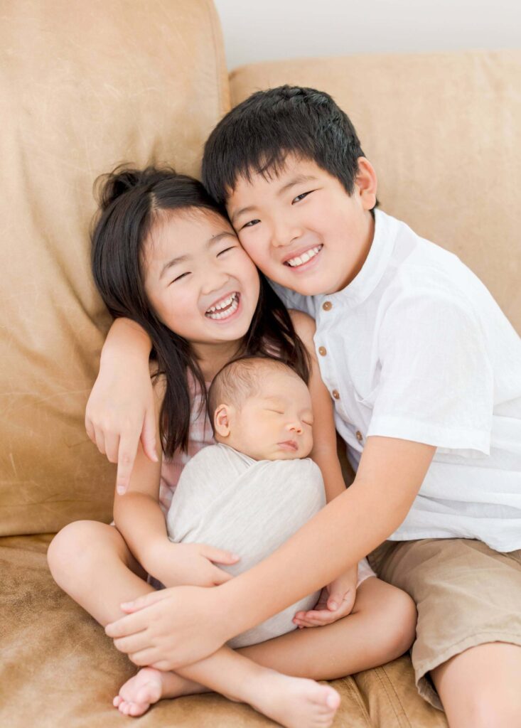 Two siblings holding their newborn family and laughing at the camera.