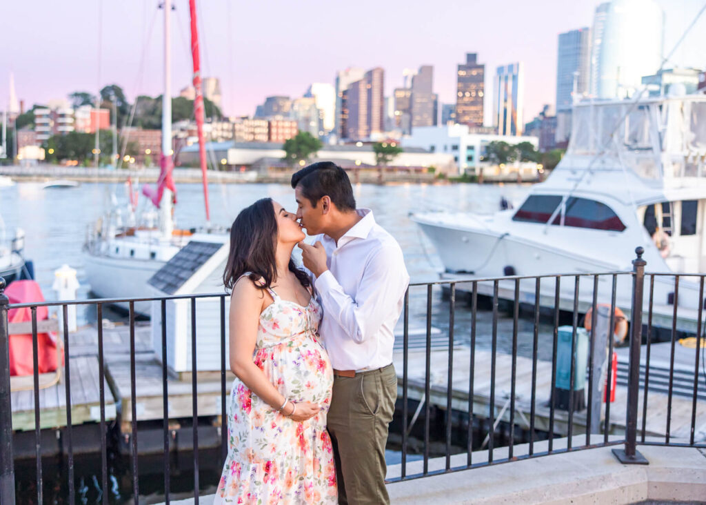 A pregnant couple kissing in the summer with the Boston waterfront in the backdrop.