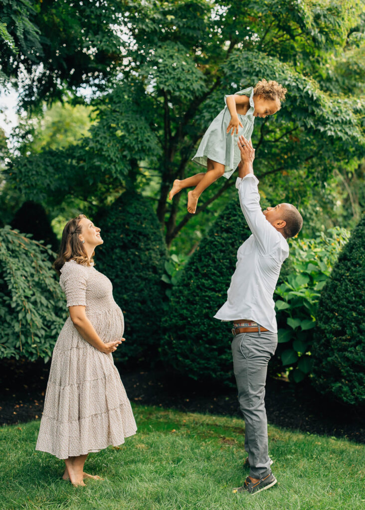An outdoor family photo with a dad throwing his daughter in the air and the pregnant mom looking and smiling. 
