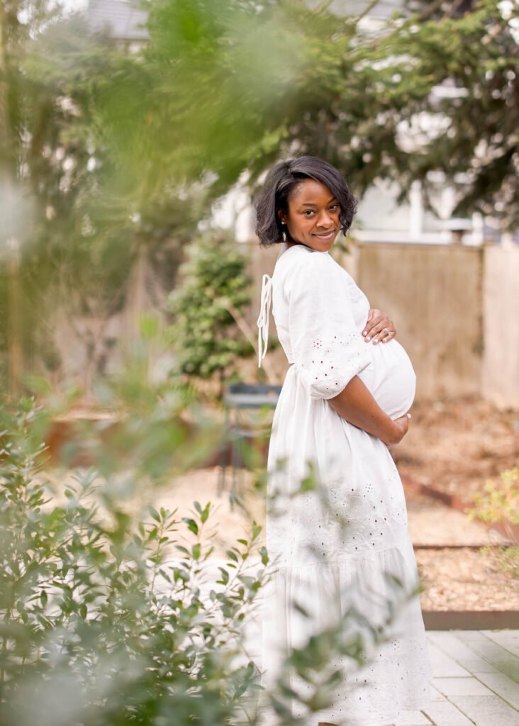 An afro American pregnant mom holding her belly and smiling at the camera with a bush in the forefront. 